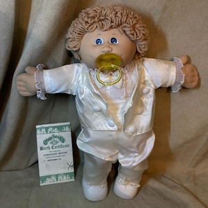 Op de afbeelding: Een Cabbage Patch Kids pop met lichtbruin garen haar en blauwe ogen, gekleed in een wit satijnen smokingjasje, bijpassende broek en een wit overhemd met een gele fopspeen in de mond. Een geboortecertificaat voor Kirkwood Bennie is inbegrepen.