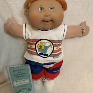 May include: A Cabbage Patch Kids doll with orange hair, green eyes, and a smiling expression. The doll wears a white and blue striped shirt with a helicopter graphic, red, white, and blue striped shorts, and white shoes. A birth certificate is included.
