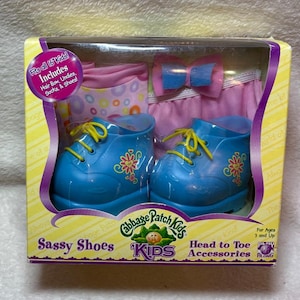 Könnte beinhalten: Ein Cabbage Patch Kids Sassy Shoes Zubehörset in einer Schachtel. Enthält blaue Schuhe mit gelben Schnürsenkeln und Blumenmustern, eine rosa Haarschleife und gemusterte Stoffartikel. Der Verpackungstext lautet Fits all 16 Kids! Includes Hair Bow, Undies, Socks, & Shoes!
