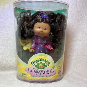 May include: A Cabbage Patch Kids Lil' Sprouts doll with dark brown curly hair in pigtails, wearing a purple polka-dot outfit with a pink belt. The doll is inside a clear cylindrical plastic package labeled Cabbage Patch Kids Lil' Sprouts.