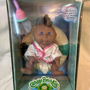 May include: A Cabbage Patch Kids Bath Baby doll in its original packaging. The doll has dark skin, blue eyes, and a tuft of dark hair. It wears a floral robe and sits on a floating bath seat. The box includes a pink and green shower accessory.