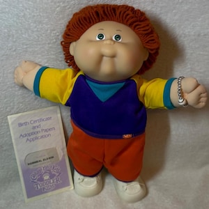 Op de afbeelding: Een Cabbage Patch Kids pop met roodbruin wollen haar en groene ogen, gekleed in een geel, paars en blauwgroen shirt met een oranje broek en witte schoenen. Een zilverkleurige kralenarmband zit om de pols, inclusief een geboortecertificaat.