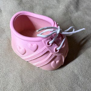 May include: A light pink ceramic planter shaped like a baby shoe, featuring embossed heart details on the sides and front. The shoe has white fabric laces tied in a bow across the top, with a hollow interior for holding small items or plants.