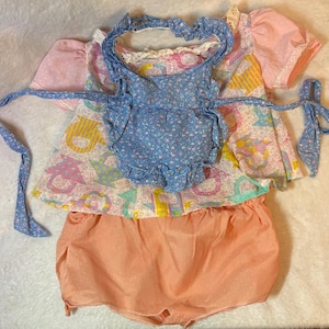 May include: A set consisting of a short-sleeved top with a white, pink, yellow, and green patterned print, a pair of solid peach-colored bloomers, and a blue floral-patterned apron with ruffled edges and long ties for securing around the waist.