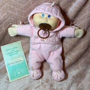 Op de afbeelding: Een Cabbage Patch Kids pop met blauwe ogen en een bruine fopspeen, gekleed in een lichtroze gebreide outfit met capuchon en kleine lintjes. Naast de pop ligt een rechthoekig document met de titel Birth Certificate and Adoption Papers Application.