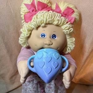 Peut inclure: Une poupée à la peau claire, aux yeux bleus et aux cheveux en laine blonde coiffés en deux couettes avec des nœuds roses. La poupée porte un haut rose clair à manches longues et tient un gobelet en plastique bleu clair avec des cœurs.