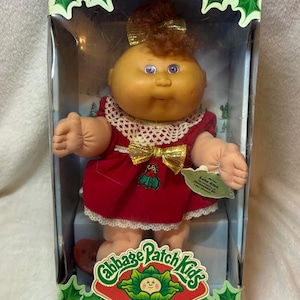 May include: A Cabbage Patch Kids Holiday Baby doll in a box. The doll has reddish-brown hair with a gold bow, purple eyes, and wears a red dress with a white lace collar and gold bow. The box features holly graphics and the text Special Edition.