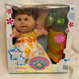 Pode incluir: Uma boneca Cabbage Patch Kids com cabelo castanho encaracolado e olhos verdes, vestindo um vestido amarelo e branco com estampa de patinhos. A boneca está numa caixa com uma mamadeira verde, uma chupeta verde e um cobertor amarelo com borda laranja.