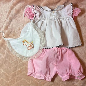 May include: A white blouse with short pink gingham sleeves, a white bib with a lace border and an embroidered orange unicorn, and pink bloomers. The blouse features a Peter Pan collar, two white buttons, and black scalloped trim edges.