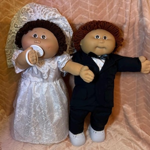 Puede incluir: Dos muñecos vestidos para una boda. El muñeco de la izquierda tiene pelo de lana marrón oscuro, un velo de encaje blanco y un vestido de satén blanco, sosteniendo un chupete blanco. El de la derecha tiene pelo de lana rojizo, traje negro.