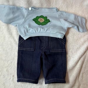 May include: A light blue doll outfit featuring a long-sleeved top and denim shorts. The top has a green cabbage applique with a smiling face. The shorts are dark blue denim with white stitching.