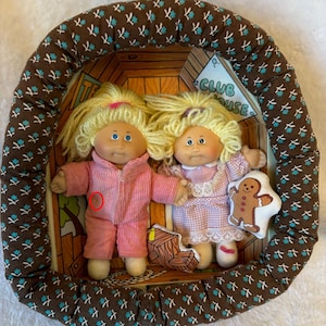 May include: Two Cabbage Patch Kids dolls in a decorative frame. One doll wears a pink jumpsuit, the other a pink and white dress. A gingerbread man plushie is held by one doll. The frame has a brown floral pattern.