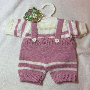 May include: A knitted outfit for a doll featuring a white and mauve striped short-sleeve top and matching mauve overalls with white horizontal stripes. The overalls have two white buttons on the front straps and a Cabbage Patch Kids tag.