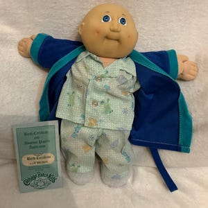 May include: A Cabbage Patch Kids doll wearing pajamas and a blue robe. The doll has blue eyes, rosy cheeks, and a bald head. The pajamas are light blue with a pattern of bunnies and other shapes. A birth certificate is included.