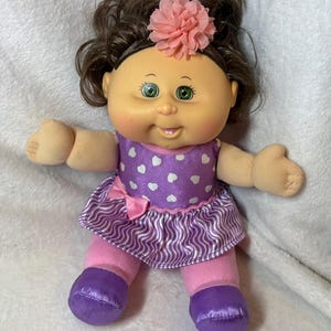 May include: A Cabbage Patch Kid doll with brown hair styled in pigtails, a pink flower hair accessory, and green eyes. The doll wears a purple dress with white hearts, a pink bow, pink leggings, and purple shoes.