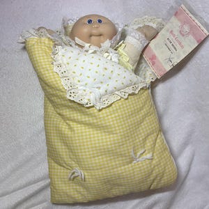May include: A Cabbage Patch Kids doll with blue eyes and a bald head, wearing a yellow and white gingham sleeping bag with white lace trim. The doll includes a birth certificate labeled Ruth Hedda, dated September 1st, with a signature by Xavier Roberts.