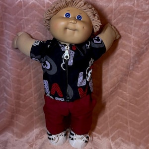 May include: A doll with light skin, blue eyes, and light brown yarn hair. It wears a black zip-up jacket featuring a white, gray, and red heart and letter pattern, solid dark red pants, and white sneakers with black stripes and white laces.