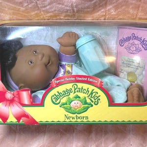 Könnte beinhalten: Eine Cabbage Patch Kids Newborn Puppe in einer gelben Schachtel mit roter Schleifengrafik. Die Puppe hat dunkle Haut, schwarzes Haar, braune Augen und trägt ein hellblaues Outfit. Enthalten sind eine hellblaue Flasche und eine rosa Karte mit dem Text Cabbage Patch Kids.
