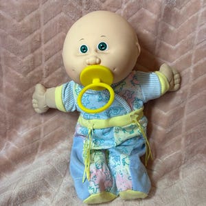 Könnte beinhalten: Eine glatzköpfige Babypuppe mit rundem Kopf, grünen Augen und Grübchen in den Wangen. Die Puppe hat einen leuchtend gelben Schnuller im Mund und trägt einen hellblau-weiß gemusterten Strampler mit kleinen Stern- und Tiermotiven.