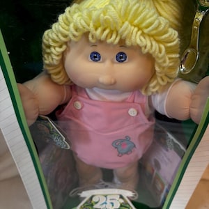 May include: A Cabbage Patch Kids doll with yellow yarn hair and blue eyes, wearing a pink overall outfit with a small elephant graphic. The doll is inside a box featuring the text 25 Cabbage Patch Kids Anniversary and includes a silver spoon.