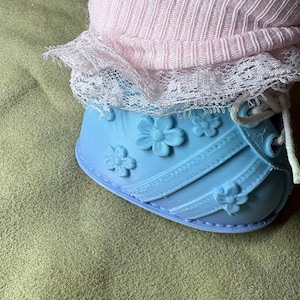 May include: A light blue molded shoe featuring raised floral details and diagonal straps, paired with a pale pink ribbed fabric cuff trimmed with white lace. The shoe includes white laces and a smooth, rounded toe design with a flat base.