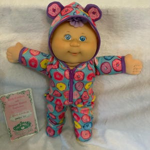 May include: A Cabbage Patch Kids doll wearing a blue onesie with a donut pattern. The onesie has a hood with purple donut-shaped ears. The doll has blue eyes and a birth certificate.