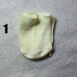 Könnte beinhalten: Eine einzelne hellgelbe Socke mit einem gerippten Bündchen am oberen Rand. Die Socke ist flach zusammengelegt und weist eine einfache Strickstruktur auf. Die Ziffer 1 ist links neben der Socke in Schwarz aufgedruckt, um sie zu kennzeichnen.