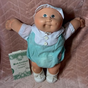 May include: A Cabbage Patch Kids doll with a round face, dimples, and green eyes, wearing a white cap, a white shirt with short sleeves, and a light turquoise romper with two white buttons. A rectangular birth certificate is placed beside the doll.