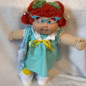 May include: A doll with bright orange yarn hair styled in pigtails with green flower accents. The doll wears light blue cat-eye glasses, a teal dress with a white collar and yellow ribbon, yellow leggings, white socks, and a white beaded bracelet.