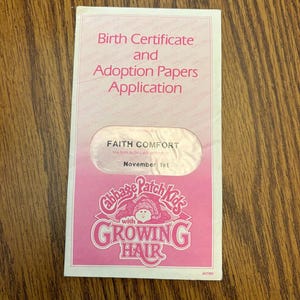 May include: A pink birth certificate and adoption papers application for a Cabbage Patch Kids doll. The certificate includes the name "FAITH COMFORT" and the birthdate "November 1st". The Cabbage Patch Kids logo with "GROWING HAIR" is at the bottom.