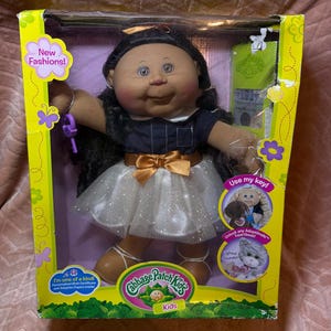Pode incluir: Uma boneca Cabbage Patch Kids de cabelo escuro e encaracolado, vestindo um top azul-marinho e uma saia branca brilhante com um laço dourado. A boneca está numa caixa amarela com o texto New Fashions, Use my key e I'm one of a kind.