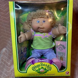 May include: A Cabbage Patch Kids doll with blonde yarn hair, blue eyes, and a light skin tone, wearing a green shirt with pink accents and light purple pants. The doll is inside a yellow and green box with the text Classic Cabbage Patch Kids.