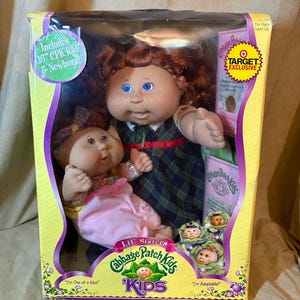 Pode incluir: Um conjunto de bonecas Cabbage Patch Kids Lil' Sisters numa caixa com uma boneca de 40,6 cm com cabelo ruivo e olhos azuis vestindo um vestido xadrez verde, e uma boneca recém-nascida menor em roupa rosa. A embalagem inclui Target Exclusive.