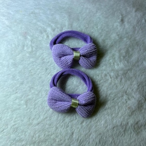 May include: Two purple hair ties featuring a textured fabric bow attached to a matching elastic band. Each bow is centred with a small, smooth, light yellow ribbon detail that secures the gathered fabric in the middle of the hair accessory.