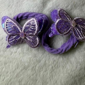 Könnte beinhalten: Zwei violette Haargummis mit schmetterlingsförmigen Verzierungen. Jeder Schmetterling hat irisierende violette Flügel mit gestickten Rändern und einen kleinen, klaren, facettierten Edelstein in der Mitte. Die Haargummis bestehen aus weichem, texturiertem Stoff.