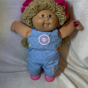 May include: A doll with light brown yarn hair styled in pigtails with bright pink bows. The doll has a round face, dimples, and wears light blue denim-style overalls featuring a repeating CPK pattern, a circular CPK logo patch, and pink shoes.
