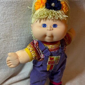 Könnte beinhalten: Eine Puppe mit heller Haut, blauen Augen und blondem Wollhaar, das mit orangefarbenen und dunkelblauen Stoffblumen verziert ist. Die Puppe trägt ein gemustertes Kurzarmshirt unter einer violetten Cordlatzhose mit einem kleinen quadratischen Aufnäher.