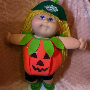 Könnte beinhalten: Eine Cabbage Patch Kids Puppe mit blondem Wollhaar und blauen Augen, bekleidet mit einem grünen Barett mit Markenlogo, einer leuchtend orangefarbenen kürbisförmigen Tunika mit schwarzem Kürbisgesicht, grünem Blattkragen, schwarzer Hose und grünen Schuhen.