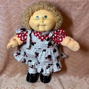 Puede incluir: Una muñeca de piel clara, ojos verdes y cabello de lana rubia rizada. Viste una camisa de lunares rojos y blancos bajo un vestido de tirantes blanco con un estampado de Minnie Mouse con siluetas negras y lazos rojos, calcetines blancos y zapatos negros.