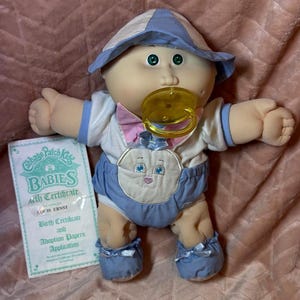 May include: A Cabbage Patch Kids baby doll with green eyes and a yellow pacifier, wearing a white and blue outfit with a bunny graphic and a matching hat. Beside the doll is a Cabbage Patch Kids Babies Birth Certificate and Adoption Papers.