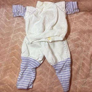 May include: A two-piece outfit consisting of a white mock neck long-sleeve top and matching pants. The top features light purple striped sleeves, while the pants have light purple striped cuffs at the bottom. The set is made of a soft fabric.