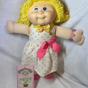 May include: A Cabbage Patch Kids doll with yellow yarn hair, blue eyes, and a cream-colored face. The doll wears a white dress with pink hearts and a pink bow. A pink box with the text "Cabbage Patch Kids" is in front of the doll.