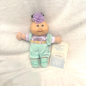 May include: A Cabbage Patch Kids doll, Elphia Cecile, with a light skin tone, blue eyes, and a lavender flower hair accessory. The doll wears a mint green outfit with a lavender collar and matching socks. A birth certificate is included.