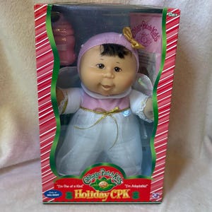 Pode incluir: Uma boneca Cabbage Patch Kids numa caixa, com um bebé de cabelo escuro, um gorro roxo claro com um laço dourado e um vestido branco com corpete rosa e fita dourada. A embalagem apresenta riscas vermelhas e brancas e o texto Holiday CPK.