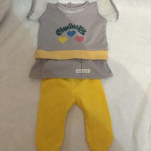 May include: A gray doll outfit featuring a short-sleeved top with the text "Cabbage Patch Kids" and three hearts. The top has white mesh sleeves and a yellow band. The outfit includes yellow pants.