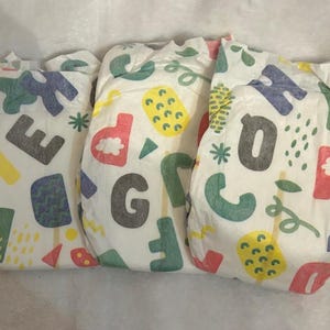 May include: Three white baby diapers with colorful letters, shapes, and floral designs. The letters are in various colors, including red, green, yellow, and gray. The diapers are arranged side by side.