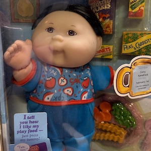May include: A Cabbage Patch Kids doll in a clear plastic package. The doll wears a blue outfit with apple graphics. The package includes miniature food items and text that reads "I tell you how I like my play food - just press my tummy!"