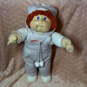 May include: A Cabbage Patch Kids doll with bright blue eyes, dimpled cheeks, and curly red yarn hair. The doll wears a light-colored knit hooded jumpsuit with white pom-poms on the drawstrings and the Cabbage Patch Kids logo in red text.
