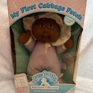 May include: A vintage Cabbage Patch Kids Babyland Collection doll in its original box. The doll has dark skin, brown eyes, and is wearing a lavender onesie and a white bonnet. The box reads "My First Cabbage Patch" and "Snuggly soft!"