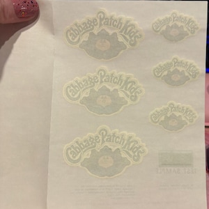 May include: A sheet of off-white adhesive stickers featuring the Cabbage Patch Kids logo in a light green hue. The sheet contains three large and three small versions of the logo, each depicting a stylized cabbage leaf design with text.